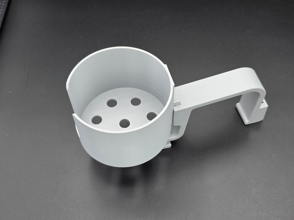 A studio-style shot of a single grey, 3D-printed cup holder attachment for a Hastings poly stock tank, isolated against a smooth black background. The holder features a deep cylindrical cup with five drainage holes at the bottom and a large side notch for mug handles. It is connected to a sturdy, integrated L-shaped mounting clip designed to hook over a thick tank rim, showcasing the smooth finish and solid plastic construction of the UV-stable PETG material.