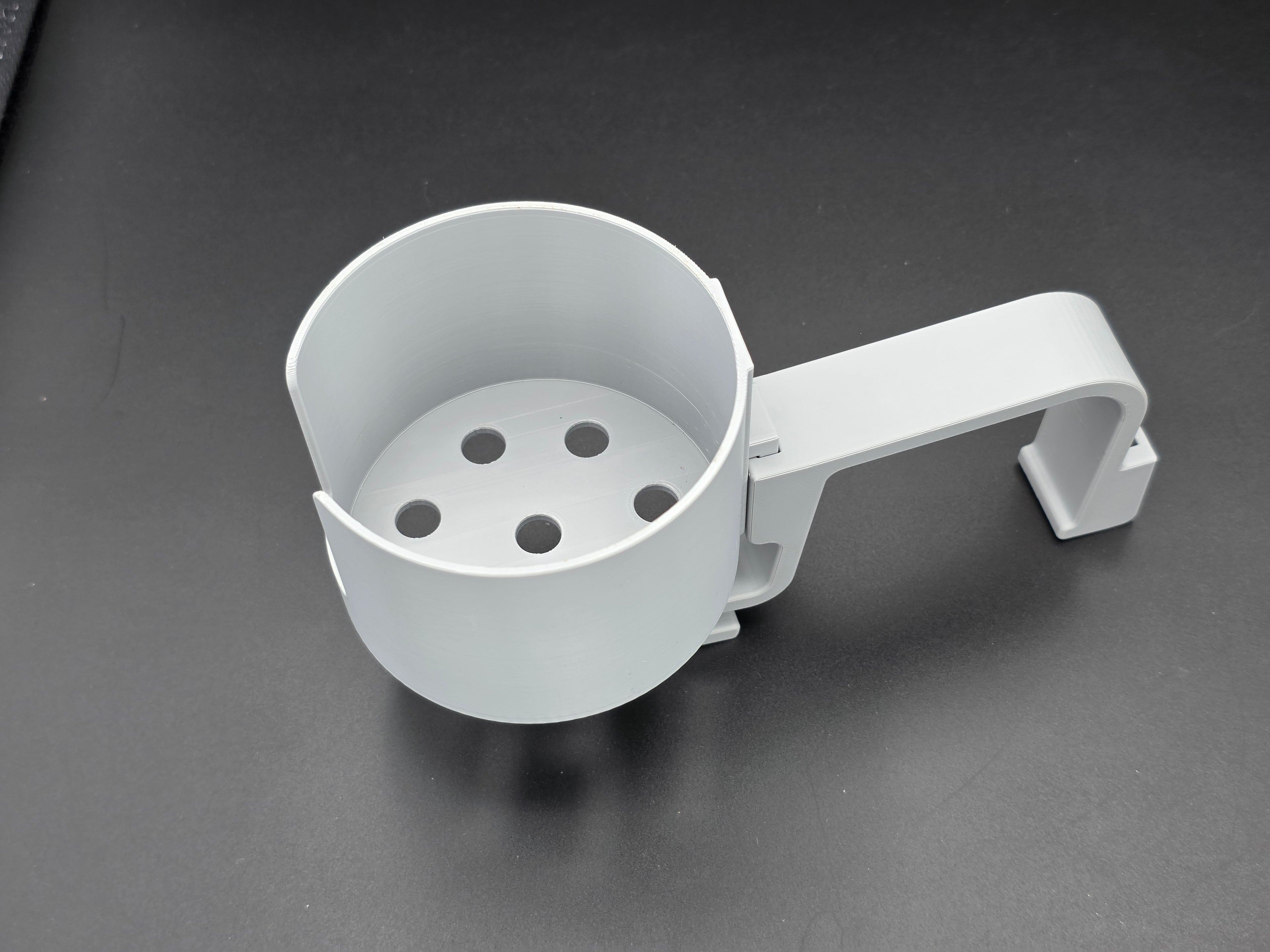 A studio-style shot of a single grey, 3D-printed cup holder attachment for a Hastings poly stock tank, isolated against a smooth black background. The holder features a deep cylindrical cup with five drainage holes at the bottom and a large side notch for mug handles. It is connected to a sturdy, integrated L-shaped mounting clip designed to hook over a thick tank rim, showcasing the smooth finish and solid plastic construction of the UV-stable PETG material.