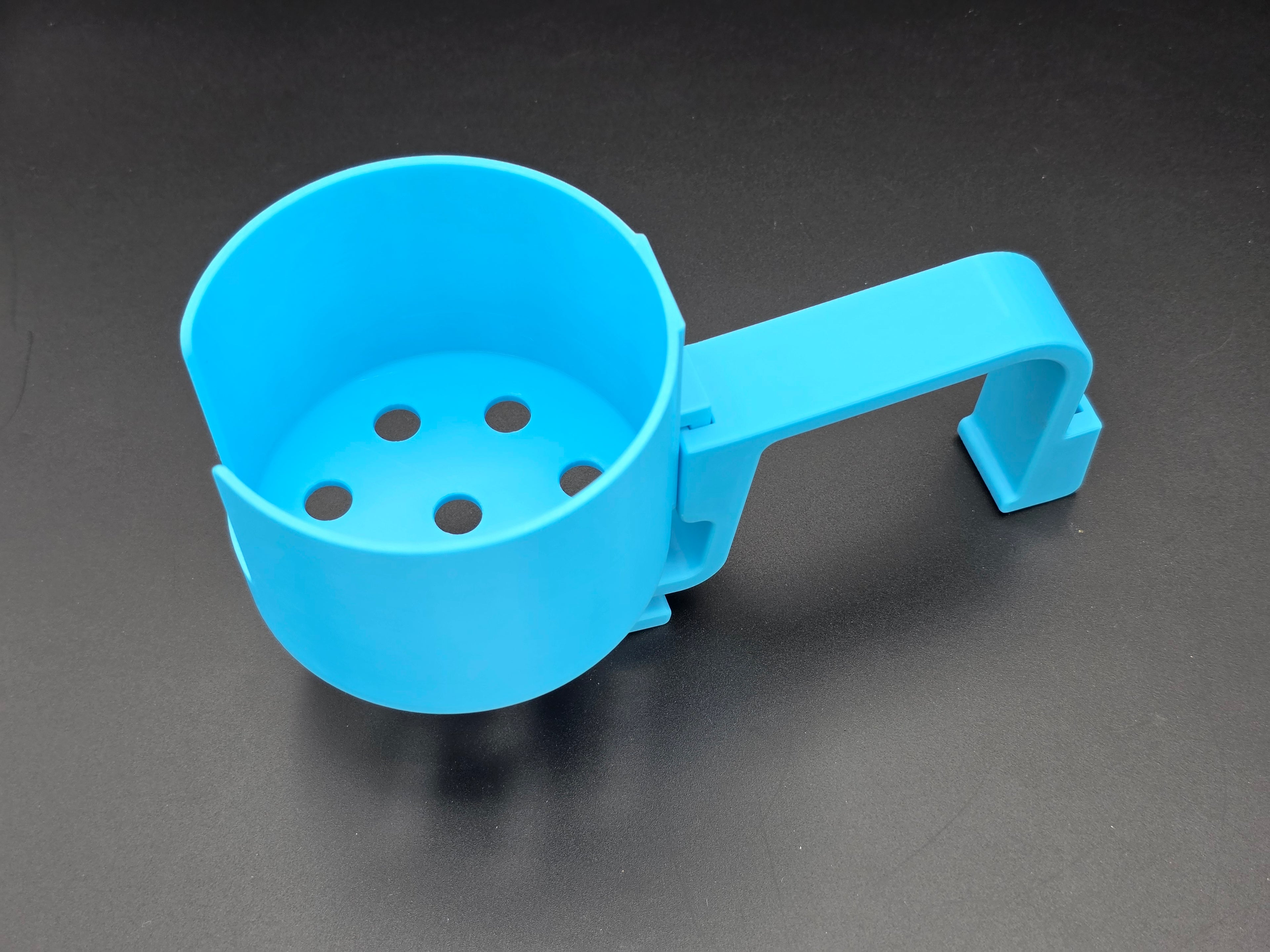 A studio-style shot of a single light blue, 3D-printed cup holder attachment for a Hastings poly stock tank, isolated against a smooth black background. The holder features a deep cylindrical cup with five drainage holes at the bottom and a large side notch for mug handles. It is connected to a sturdy, integrated L-shaped mounting clip designed to hook over a thick tank rim, showcasing the smooth finish and solid plastic construction of the UV-stable PETG material.