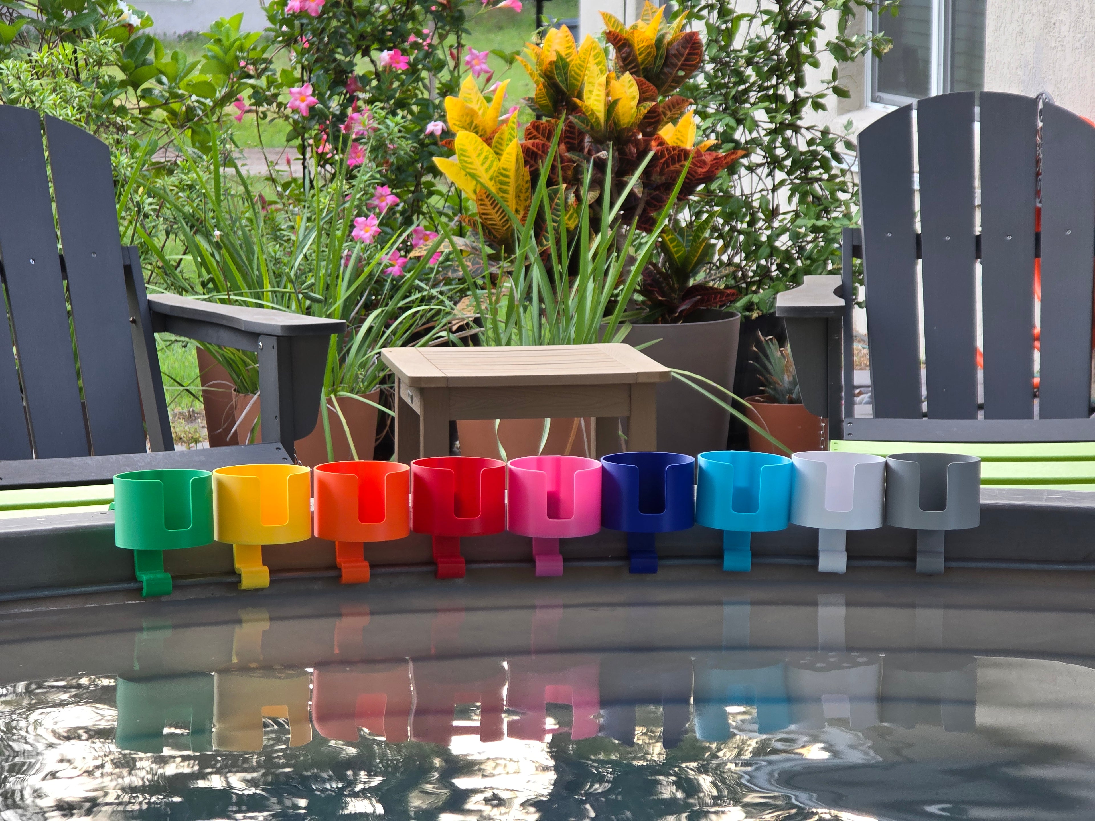 A row of colorful cup holders in various colors, including green, yellow, orange, red, pink, blue, light blue, white, and grey. Each holder is attached to the rim of a grey stock tank, creating a vibrant and organized display. The background includes outdoor furniture and lush greenery.