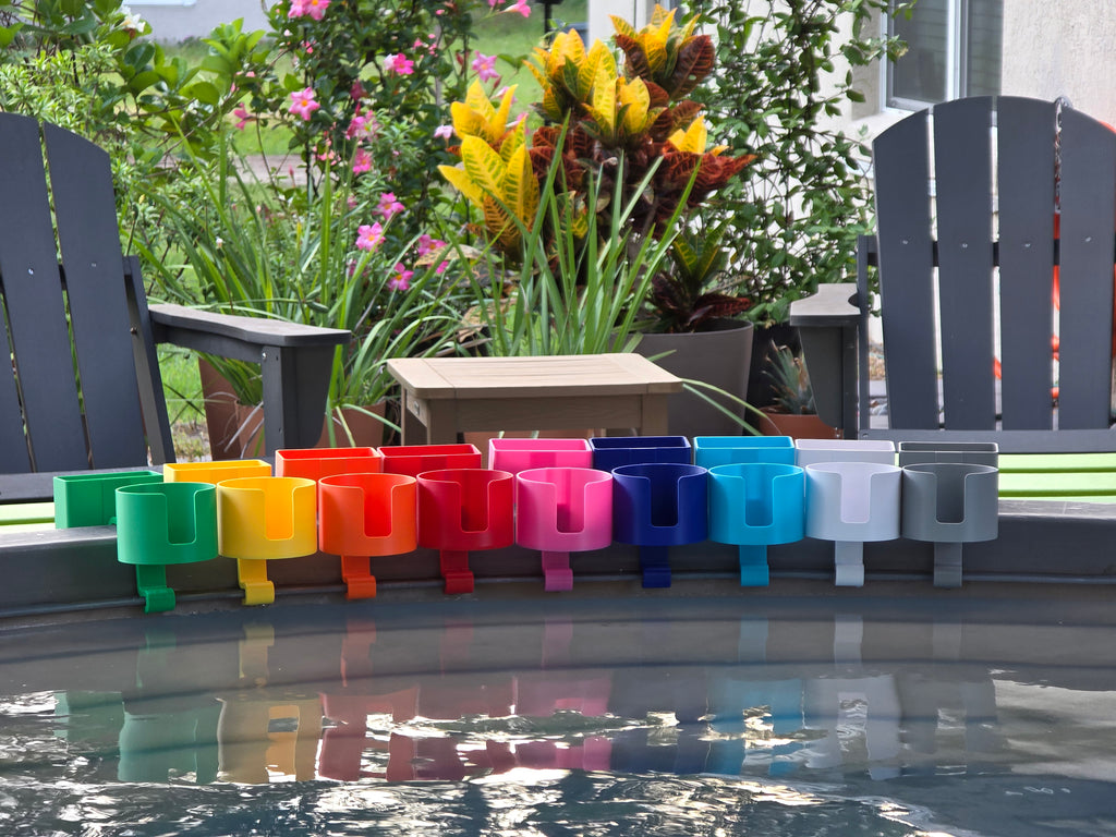 A row of colorful cup holders with cell phone caddies in various colors, including green, yellow, orange, red, pink, blue, light blue, white, and grey. Each holder is attached to the rim of a grey stock tank, creating a vibrant and organized display. The background includes outdoor furniture and lush greenery.