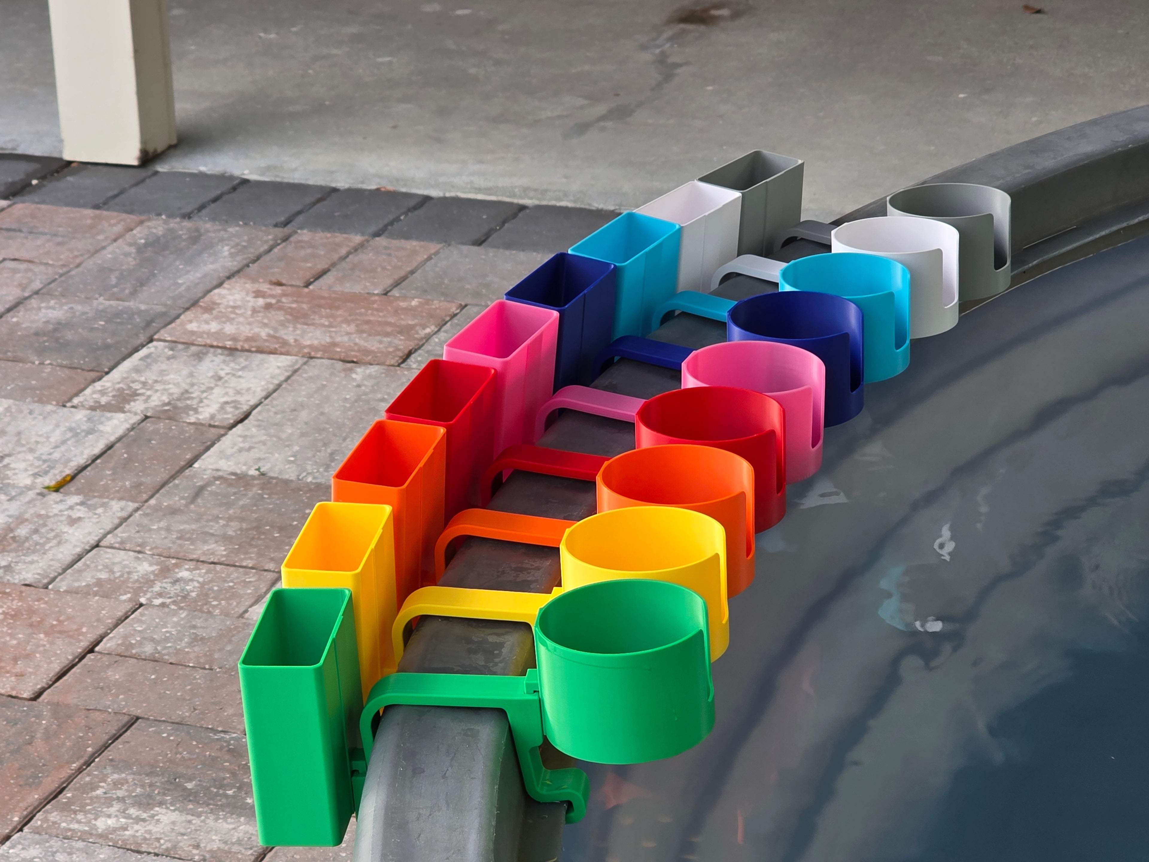 A row of colorful cup holders with cell phone caddies in various colors, including green, yellow, orange, red, pink, blue, light blue, white, and grey. Each holder is attached to the rim of a grey stock tank, creating a vibrant and organized display.