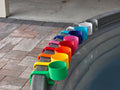 A row of colorful cup holders in various colors, including green, yellow, orange, red, pink, blue, light blue, white, and grey. Each holder is attached to the rim of a grey stock tank, creating a vibrant and organized display.