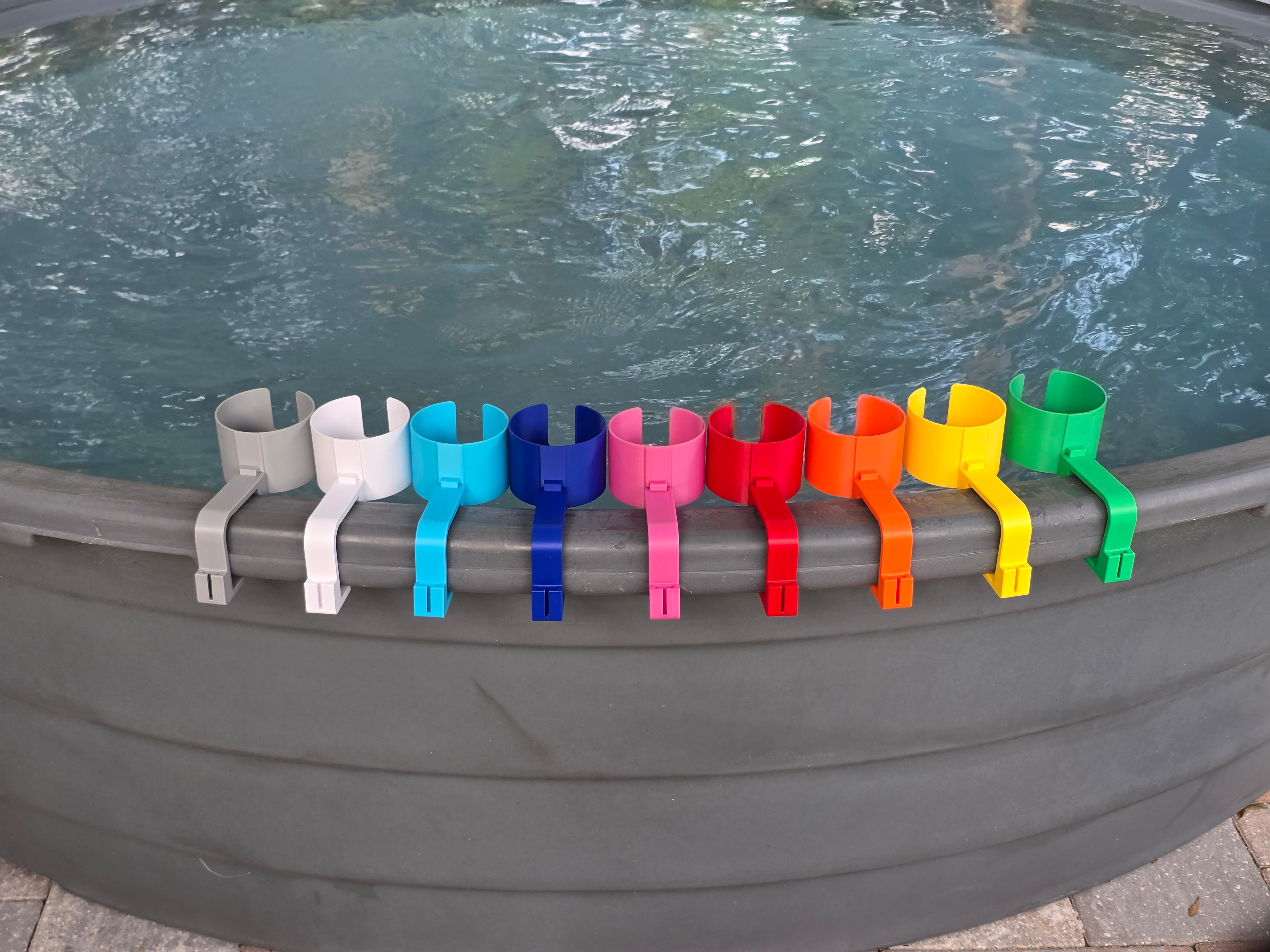 A row of colorful cup holders in various colors, including grey, white, light blue, blue, pink, red, orange, yellow, and green. Each holder has a curved design to hold a cup and a clip to attach to the edge of a stock tank. This image shows how the accessory attachment point on the outside edge of the clip is unpopulated as no cell phone holders are included in this listing. 