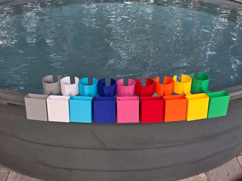 A row of colorful cup holders with cell phone caddies in various colors, including grey, white, light blue, blue, pink, red, orange, yellow, and green. Each holder has a curved design to hold a cup and a clip to attach to the edge of a stock tank. This image shows how the accessory attachment point on the outside edge of the clip is populated with cell phone holders as they are included in this listing.