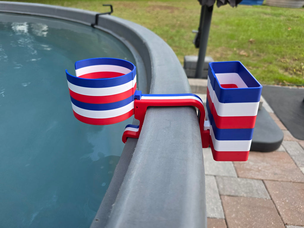 A red, white, and blue striped cup holder with an attached cell phone holder is clipped onto the edge of a stock tank. The holder is made of plastic and features a patriotic color scheme.