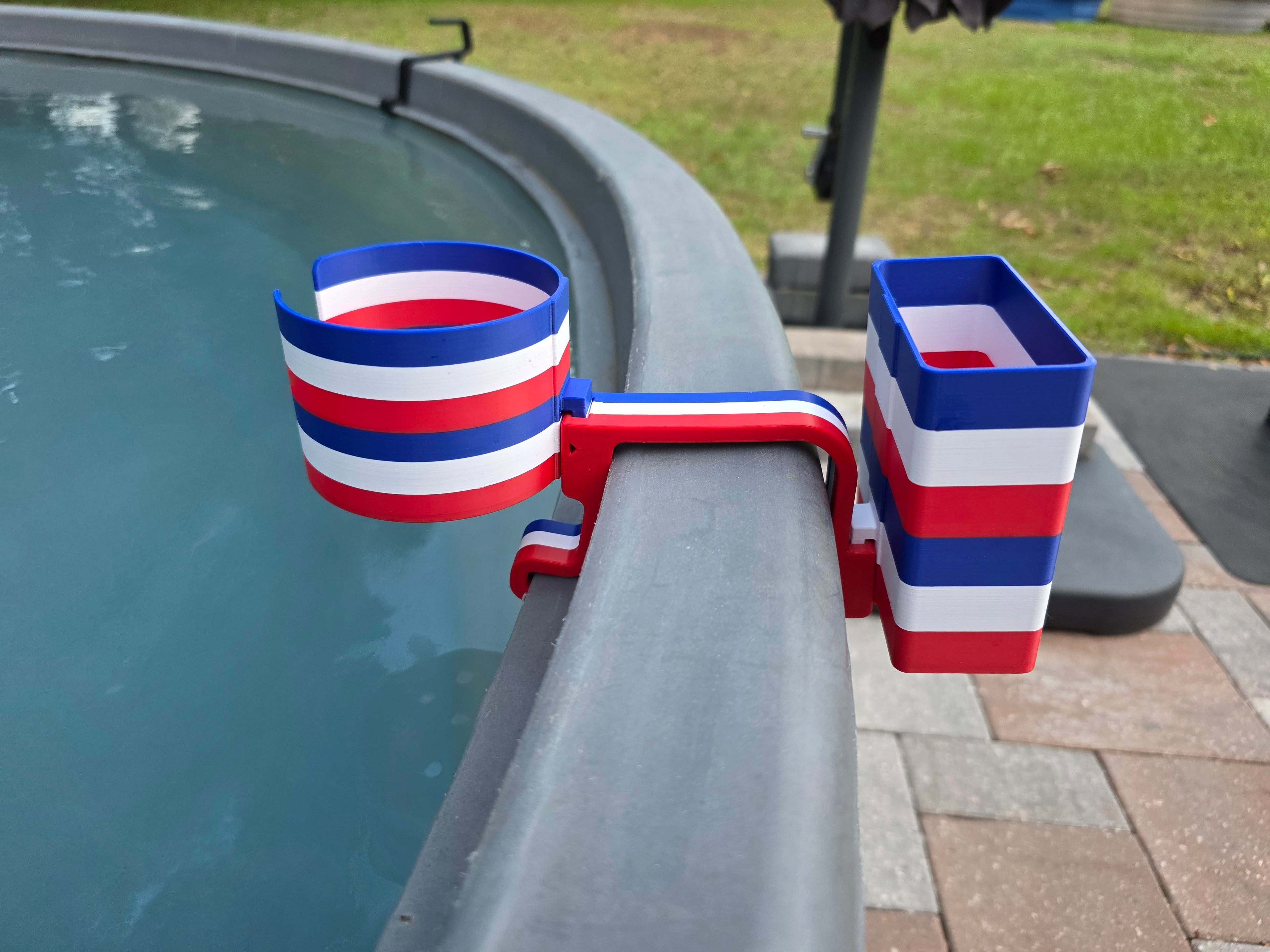 A red, white, and blue striped cup holder with an attached cell phone holder is clipped onto the edge of a stock tank. The holder is made of plastic and features a patriotic color scheme.