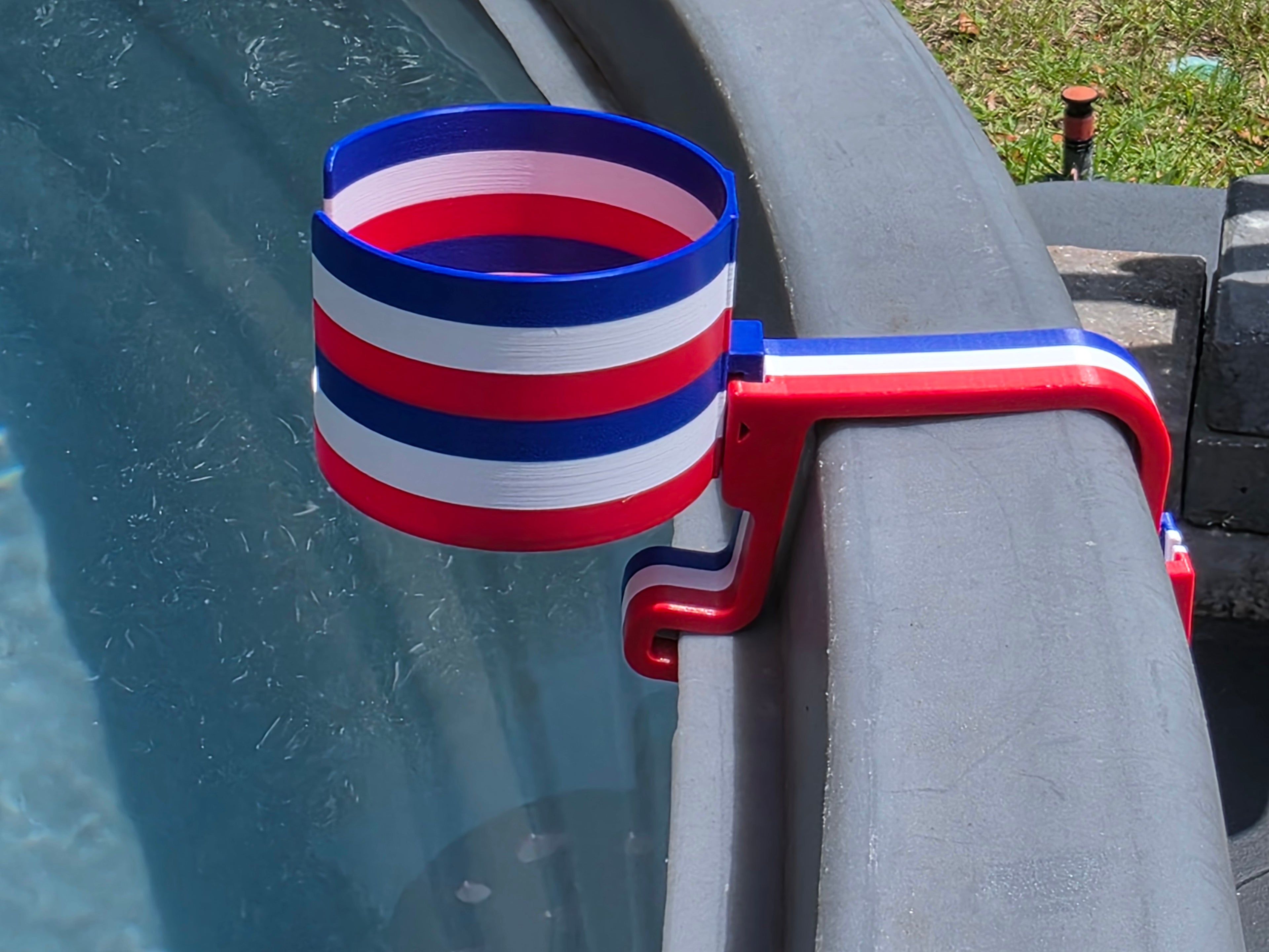 A red, white, and blue striped cup holder, clipped onto the edge of a stock tank. The holder is made of plastic and features a patriotic color scheme.
