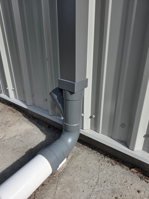 A square grey metal gutter is being adapted to 4" Sewer & Drain (S&D) PVC piping. The 3D Printed adapter and the PVC have been painted tom closely match the grey of the downspout. 