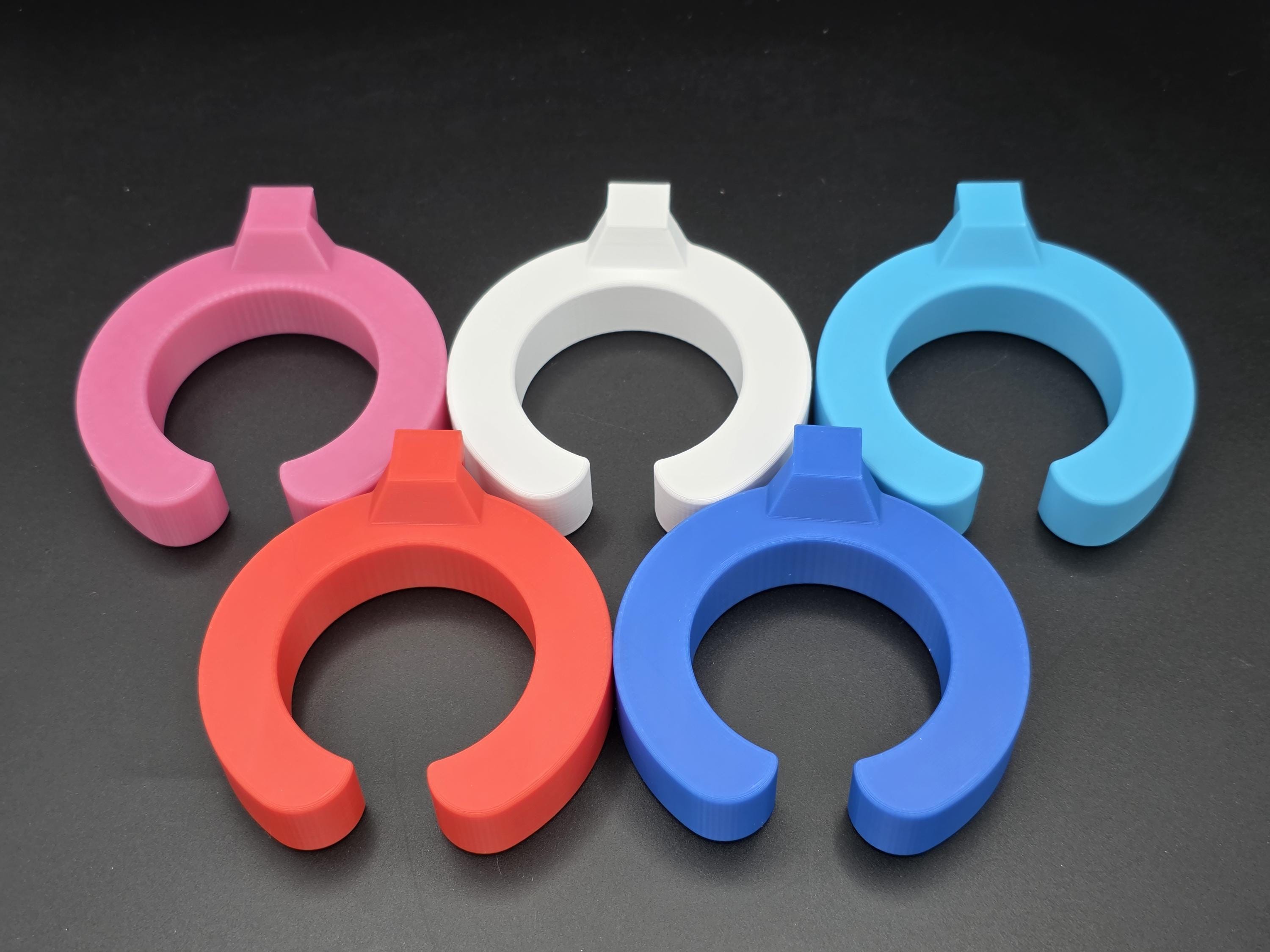 A studio shot of five colorful, C-shaped 3D-printed wine glass holders arranged on a black background. The holders are shown in pink, white, light blue, red, and blue, featuring a secure open-ring design with a mounting point at the top to slide into a stock tank bracket system.