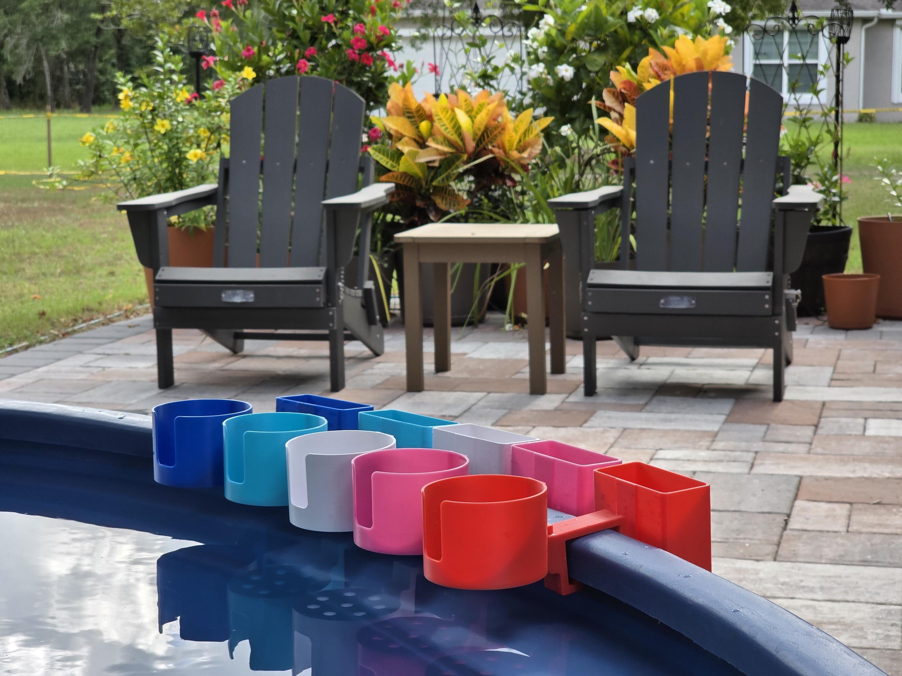 A lineup of vibrant, 3D-printed pool accessories mounted on the blue rim of a stock tank. The shot shows several colorful cup and phone holder attachments in blue, light blue, white, pink, and red, illustrating the modular clip system and the high-quality PETG finish in a backyard poolside setting.