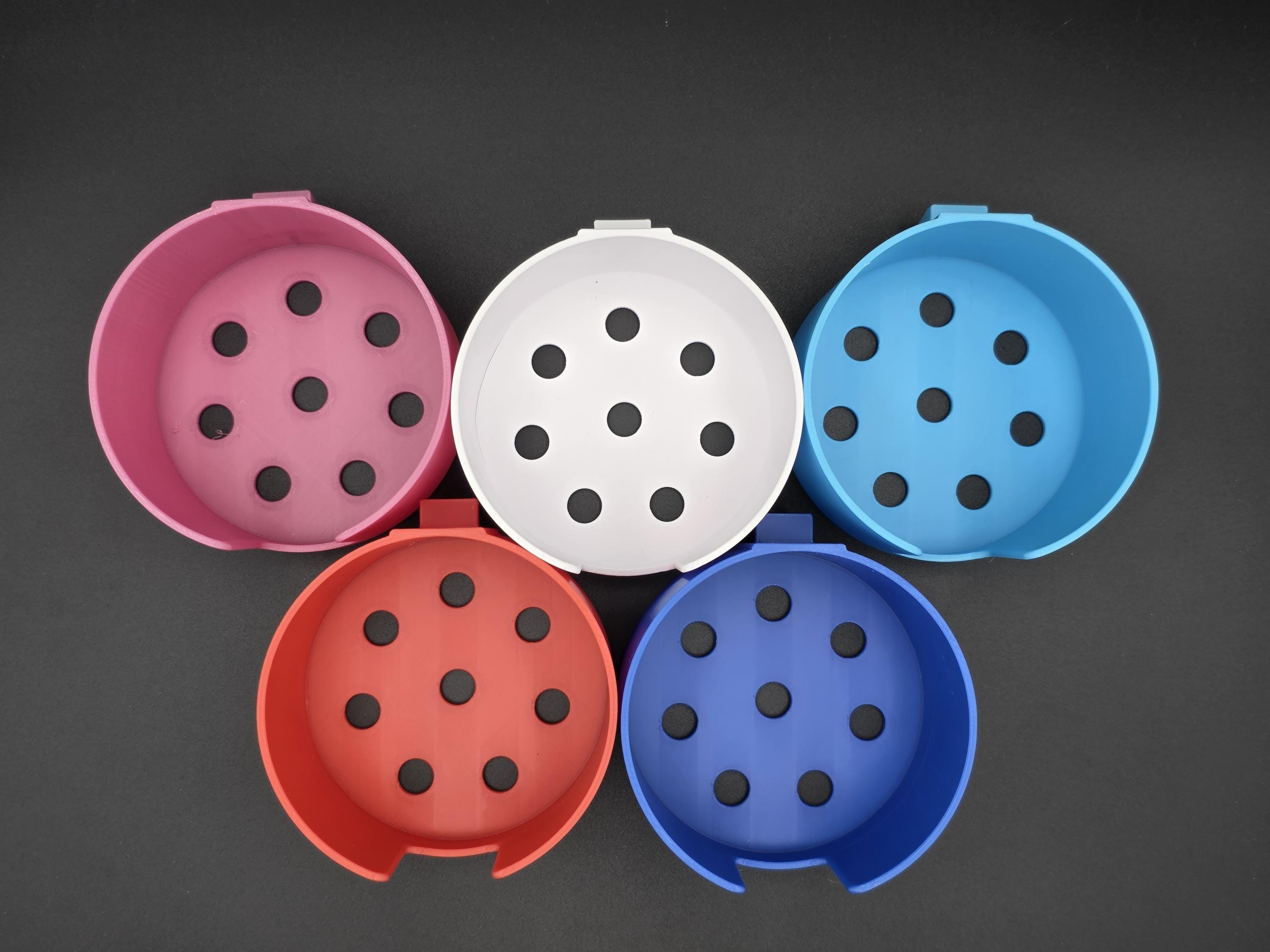 An overhead studio shot of five colorful, 3D-printed cup holders arranged on a black background. The holders are shown in pink, white, light blue, red, and blue, featuring a cutout for mug handles. 