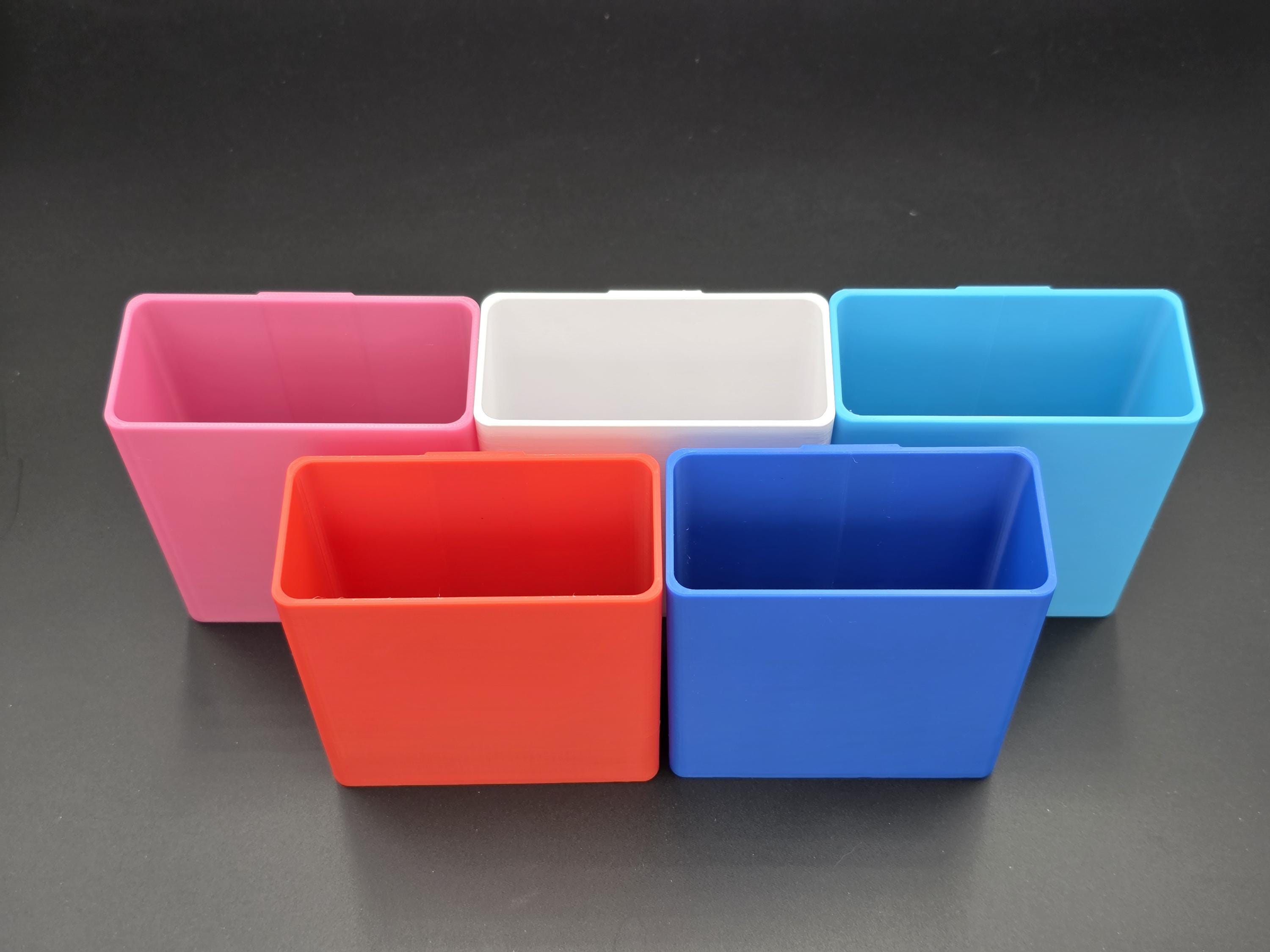 A studio shot of five colorful, 3D-printed cell phone holders arranged on a black background. The holders are shown in pink, white, light blue, red, and blue. 