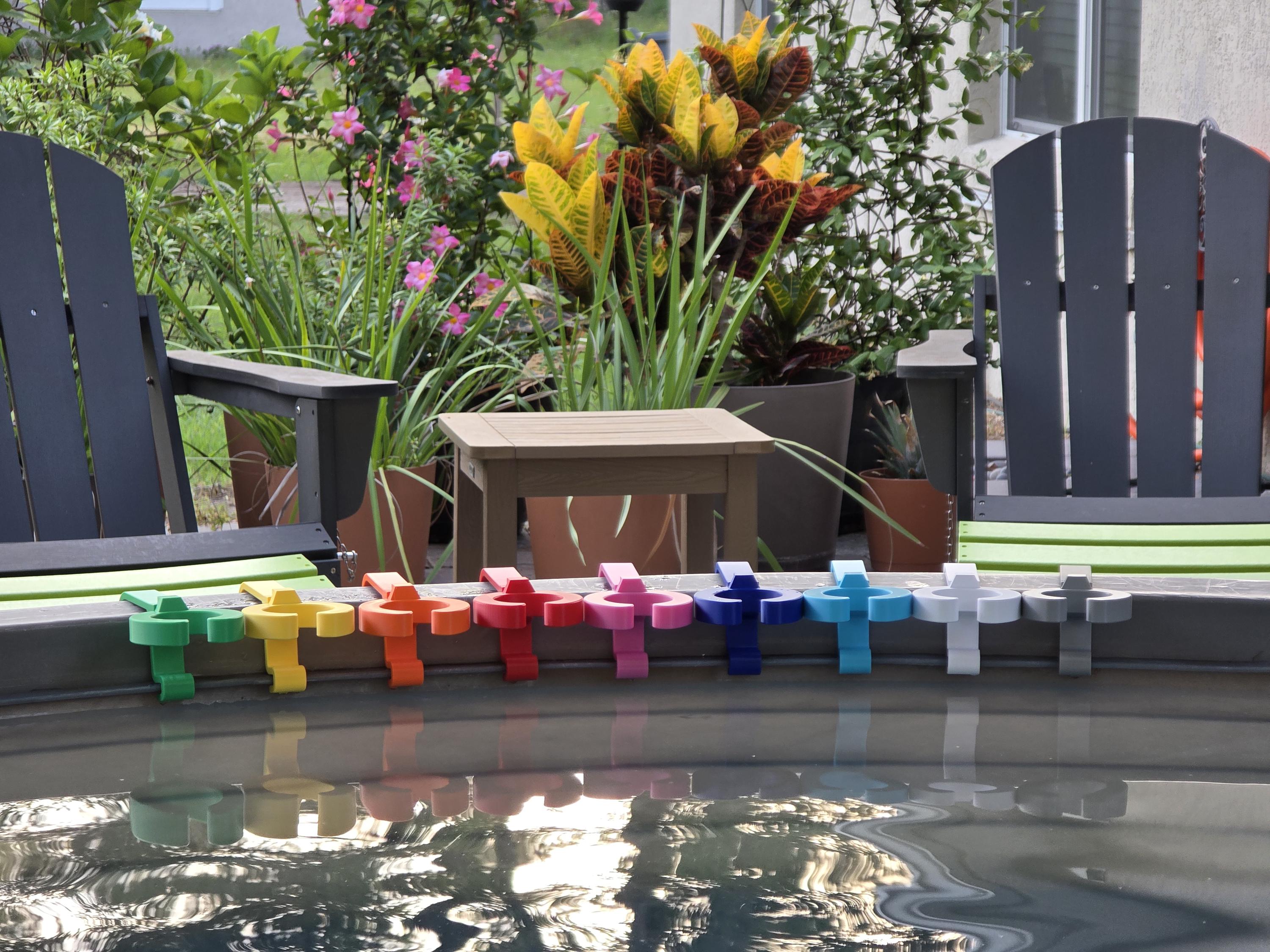 A row of colorful wine glass holders in various colors, including green, yellow, orange, red, pink, blue, light blue, white, and grey. Each holder is attached to the rim of a grey stock tank, creating a vibrant and organized display. The background includes outdoor furniture and lush greenery.