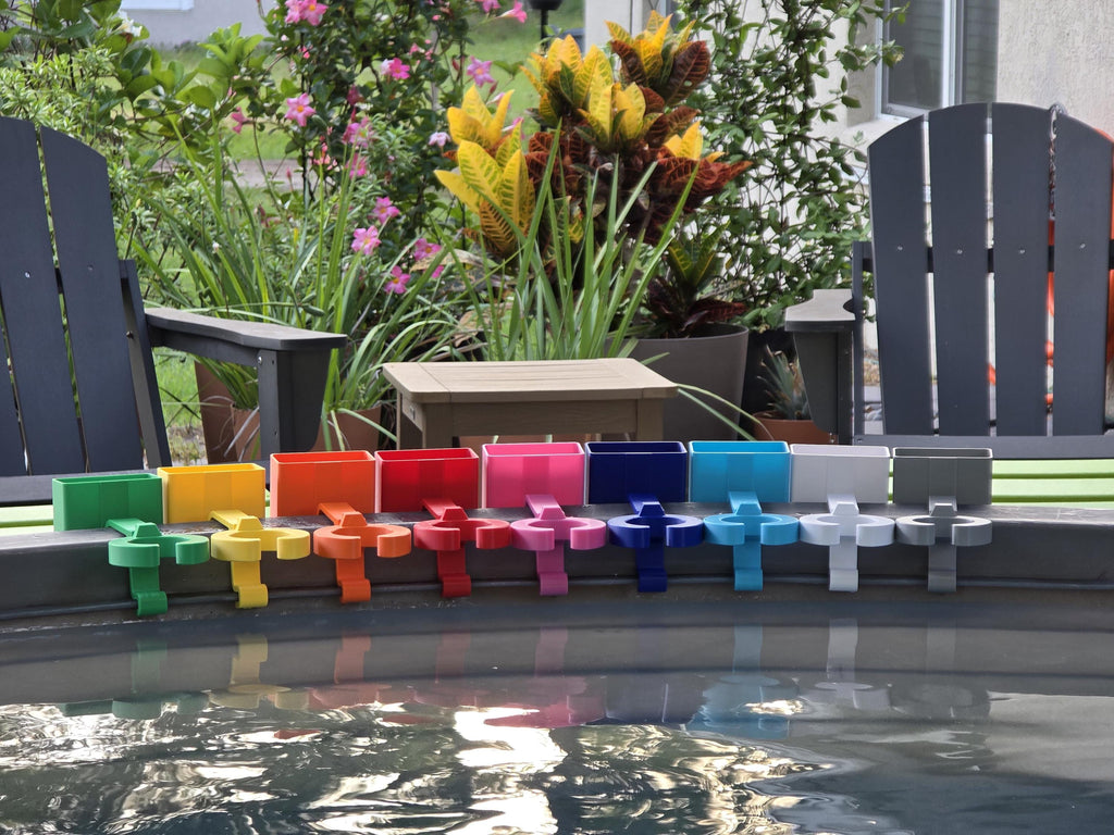 A row of colorful wine glass holders with cell phone caddies in various colors, including green, yellow, orange, red, pink, blue, light blue, white, and grey. Each holder is attached to the rim of a grey stock tank, creating a vibrant and organized display. The background includes outdoor furniture and lush greenery.