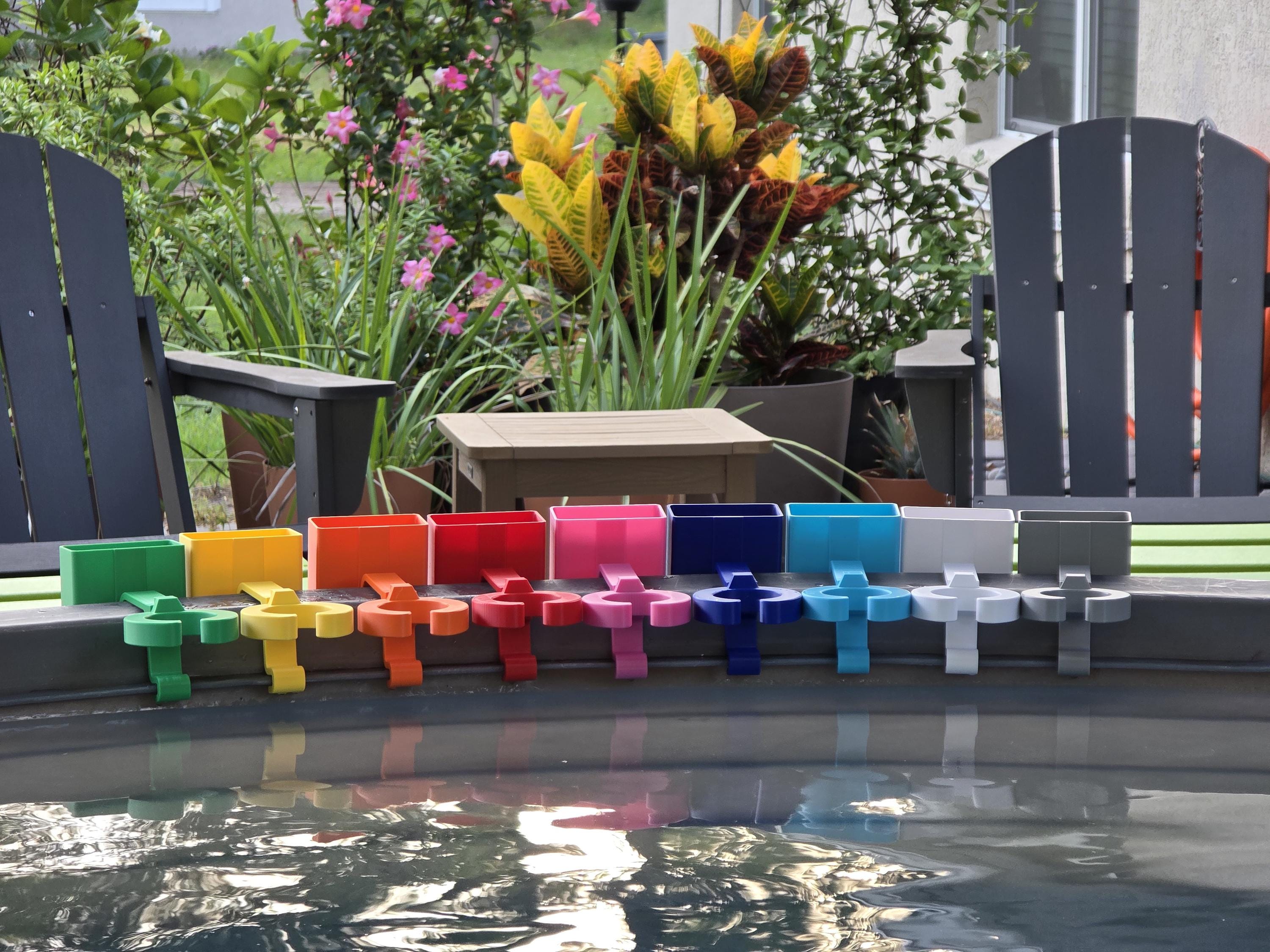 A row of colorful wine glass holders with cell phone caddies in various colors, including green, yellow, orange, red, pink, blue, light blue, white, and grey. Each holder is attached to the rim of a grey stock tank, creating a vibrant and organized display. The background includes outdoor furniture and lush greenery.