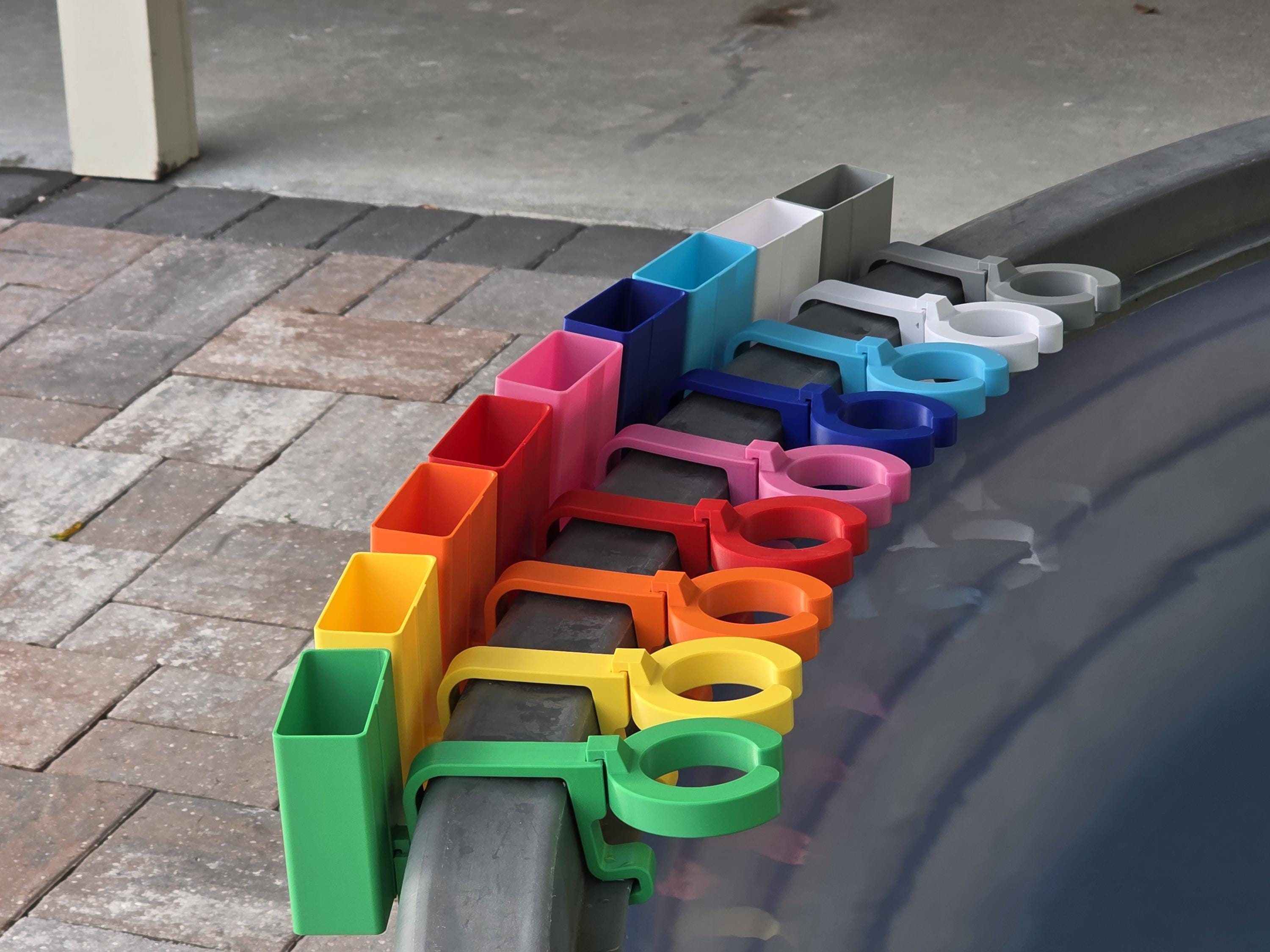 A row of colorful wine glass holders with cell phone caddies in various colors, including green, yellow, orange, red, pink, blue, light blue, white, and grey. Each holder is attached to the rim of a grey stock tank, creating a vibrant and organized display.
