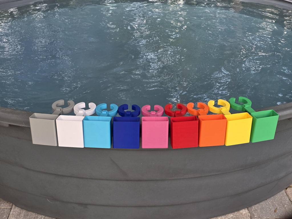 A row of colorful wine glass holders with cell phone caddies in various colors, including grey, white, light blue, blue, pink, red, orange, yellow, and green. Each holder has a curved design to hold a cup and a clip to attach to the edge of a stock tank. This image shows how the accessory attachment point on the outside edge of the clip is populated with cell phone holders as they are included in this listing.