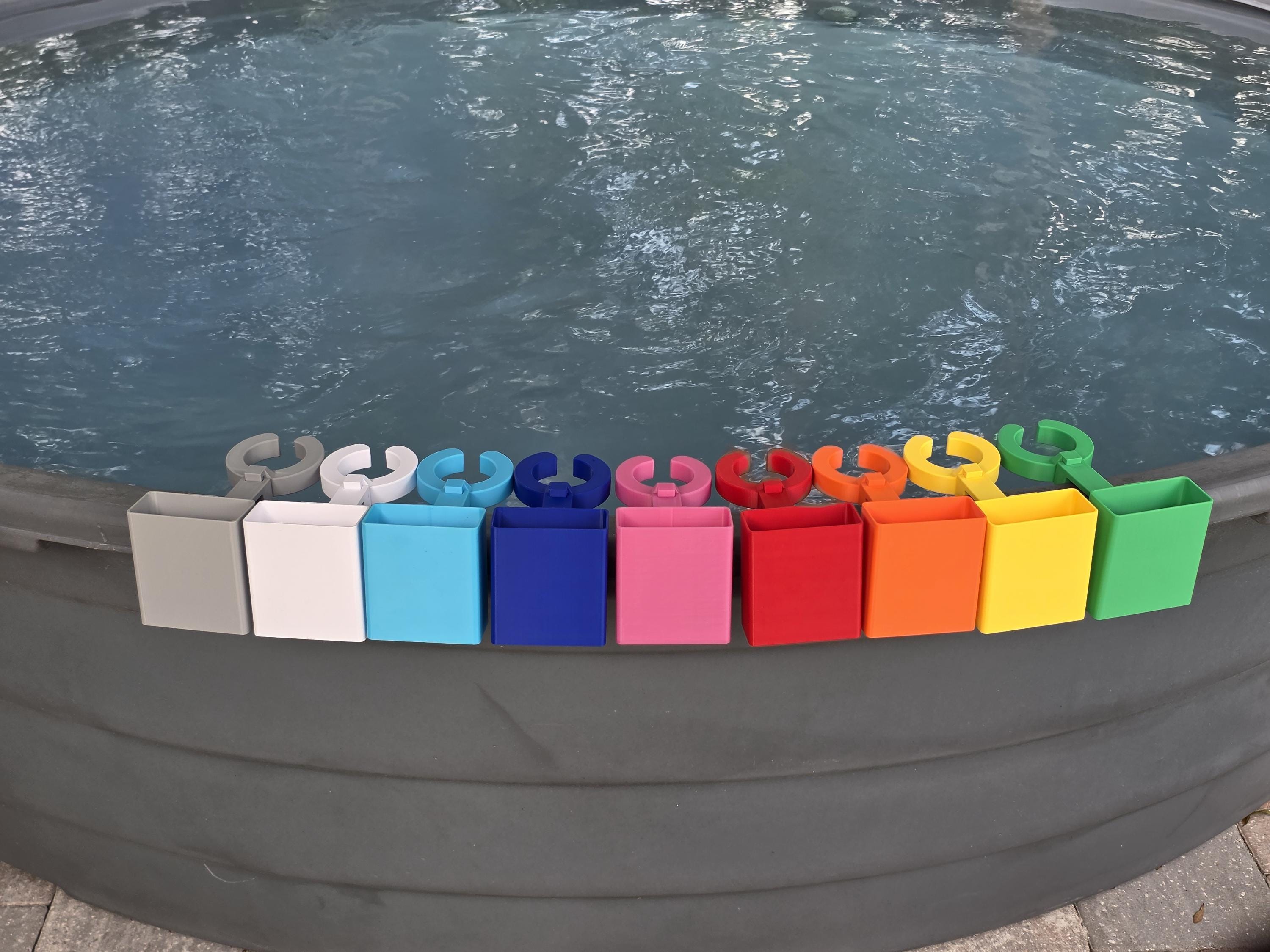 A row of colorful wine glass holders with cell phone caddies in various colors, including grey, white, light blue, blue, pink, red, orange, yellow, and green. Each holder has a curved design to hold a cup and a clip to attach to the edge of a stock tank. This image shows how the accessory attachment point on the outside edge of the clip is populated with cell phone holders as they are included in this listing.