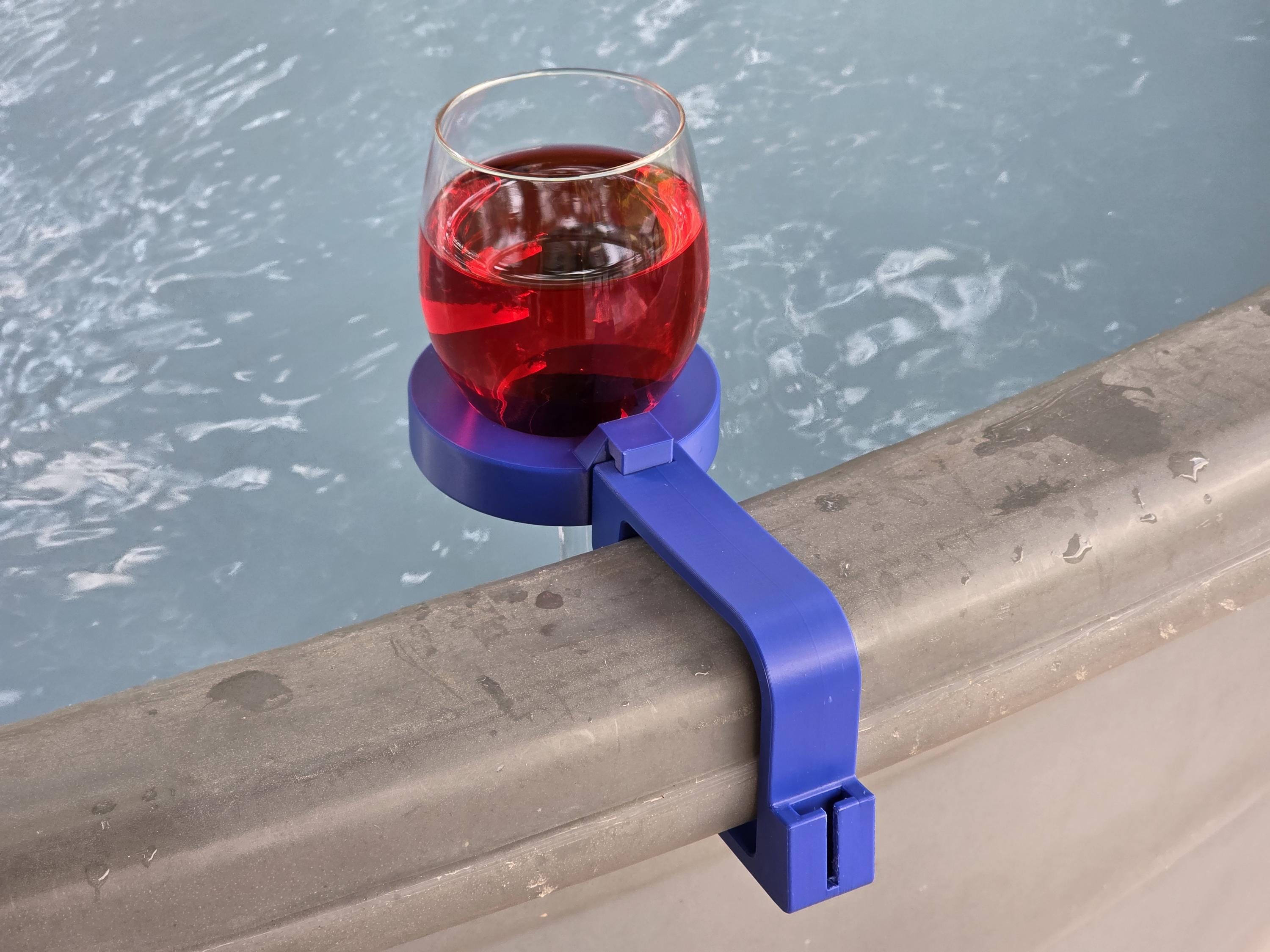A clear wine glass filled with red liquid sits in a blue wine glass holder clamped to the edge of a stock tank.
