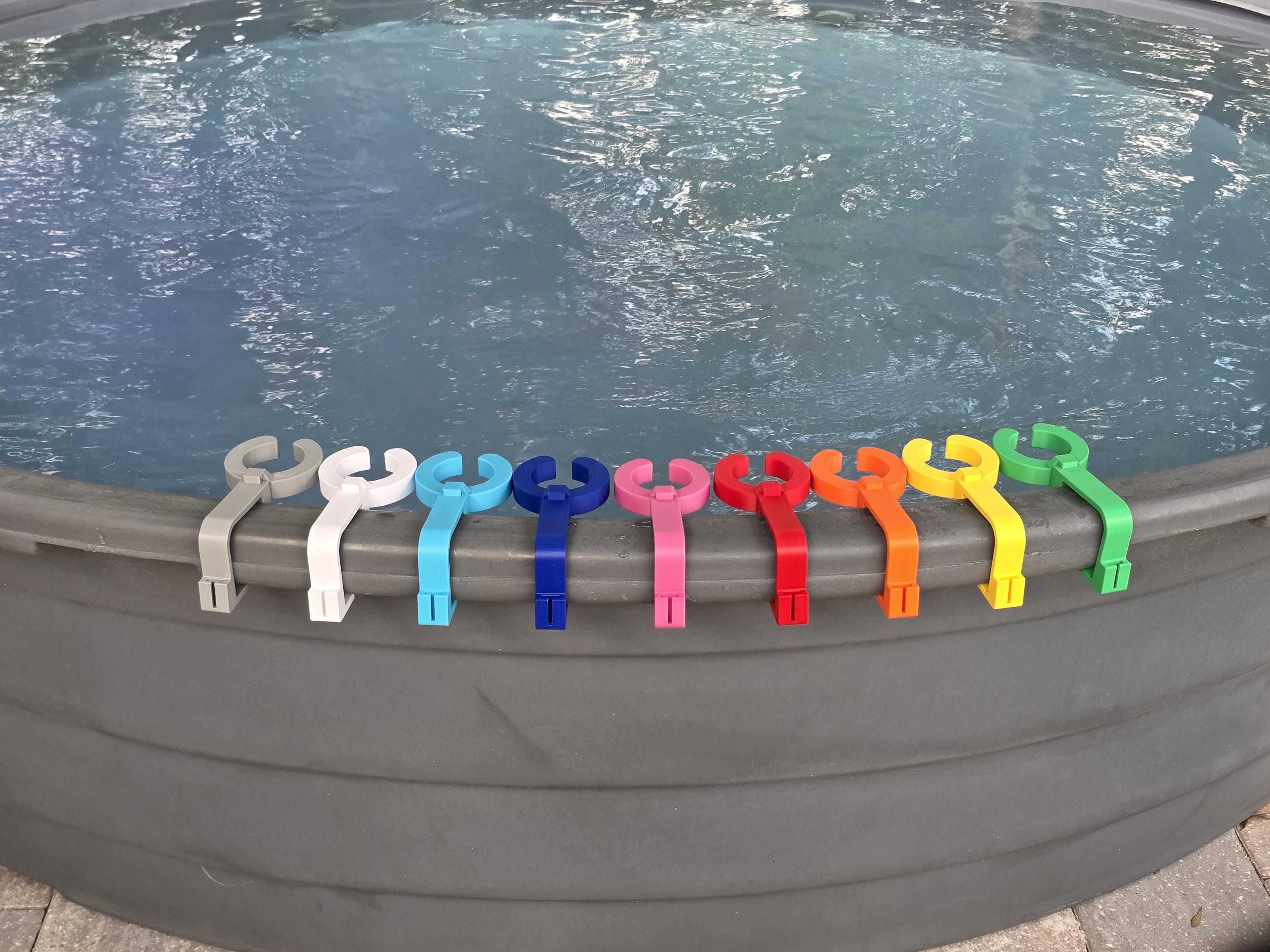 A row of colorful 3D-printed wine glass holder attachments mounted on the curved grey rim of a stock tank pool. The attachments are lined up in a vibrant spectrum of PETG plastic colors, showing the open slide-in track designed specifically to hold stemmed glassware securely. This image shows how the accessory attachment point on the outside edge of the clip is unpopulated as no cell phone holders are included in this listing.