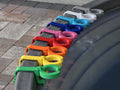 A row of colorful 3D-printed wine glass holder attachments mounted on the curved grey rim of a stock tank pool. The attachments are lined up in a vibrant spectrum of PETG plastic colors, showing the open slide-in track designed specifically to hold stemmed glassware securely.