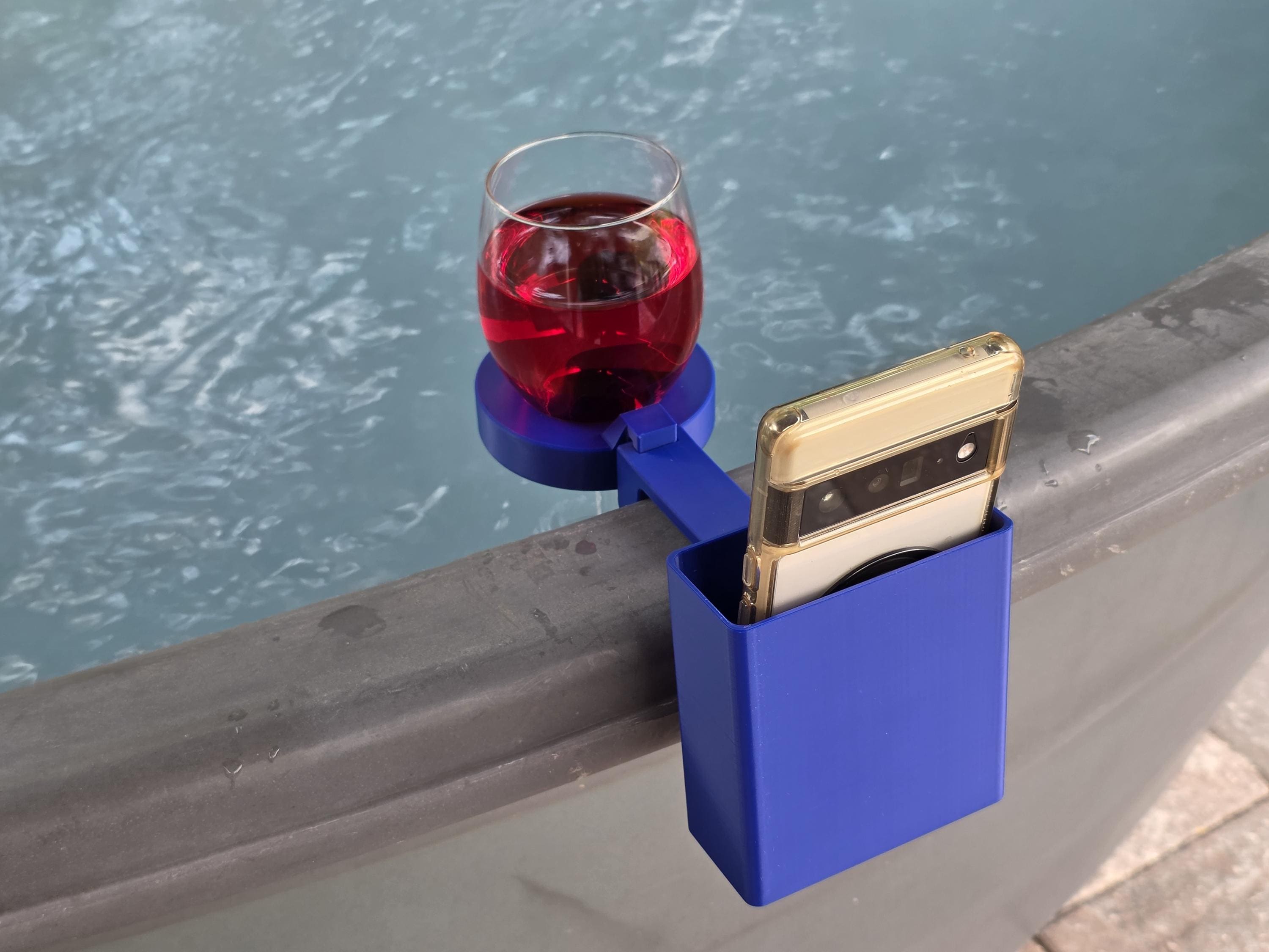 A clear wine glass filled with red liquid and sits in a blue wine glass holder along with a white cell phone in a blue cell phone pocket and both are clipped to the edge of a stock tank.